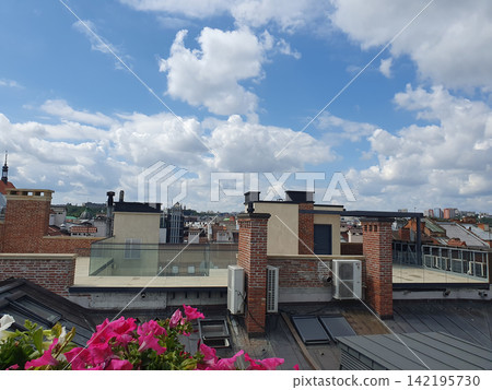 Modern roof terrace with pink petunias, brick chimneys and air conditioners. View of blue sky with snow-white clouds Modern roof terrace with pink petunias, brick chimneys and air conditioners. View of blue sky with snow-white clouds 142195730