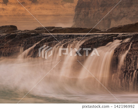 Long exposure photography of silky smooth water cascading over rocky cliff terraces at Klayar Beach (Pantai Klayar) during warm golden hour sunset, Pacitan, East Java, Indonesia. Long exposure photography of silky smooth water cascading over rocky cliff terraces at Klayar Beach (Pantai Klayar) during warm golden hour sunset, Pacitan, East Java, Indonesia. 142196992