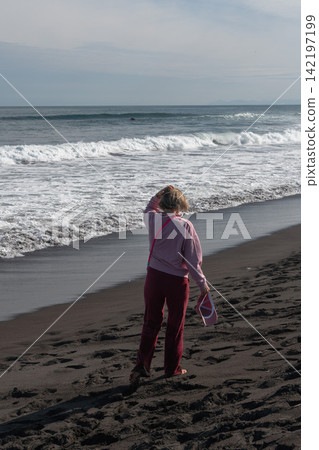Woman stands facing Pacific Ocean waves on black volcanic shore at Khalaktyrsky Beach, Kamchatka. Travel, nature tourism, coastal lifestyle, contemplation, solitude, summer vacation. Woman stands facing Pacific Ocean waves on black volcanic shore at Khalaktyrsky Beach, Kamchatka. Travel, nature tourism, coastal lifestyle, contemplation, solitude, summer vacation. 142197199