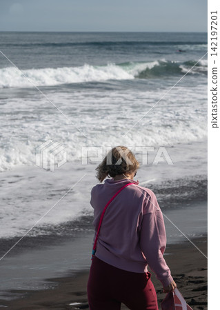 Woman watches breaking Pacific waves from black volcanic beach at Khalaktyrsky Beach, Kamchatka. Travel, coastal lifestyle, nature tourism, contemplation, solitude, summer vacation. Woman watches breaking Pacific waves from black volcanic beach at Khalaktyrsky Beach, Kamchatka. Travel, coastal lifestyle, nature tourism, contemplation, solitude, summer vacation. 142197201