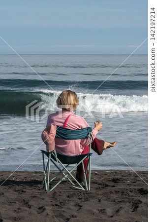 Woman relaxes by Pacific Ocean with glass of champagne while seated on black volcanic sand in Kamchatka. Travel, coastal recreation, peaceful lifestyle, outdoor leisure, solitude, summer vacation. Woman relaxes by Pacific Ocean with glass of champagne while seated on black volcanic sand in Kamchatka. Travel, coastal recreation, peaceful lifestyle, outdoor leisure, solitude, summer vacation. 142197214
