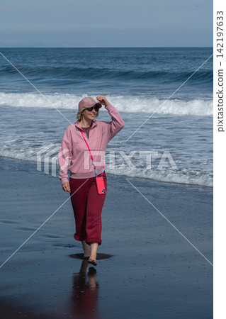 Woman in pink clothes stands barefoot on wet black volcanic sand beside Pacific Ocean waves in Kamchatka. Travel, coastal lifestyle, relaxation, nature tourism, freedom, summer vacation. Woman in pink clothes stands barefoot on wet black volcanic sand beside Pacific Ocean waves in Kamchatka. Travel, coastal lifestyle, relaxation, nature tourism, freedom, summer vacation. 142197633
