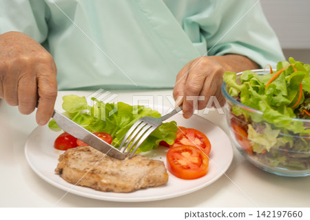 Asian elderly woman patient eating pork chop stake and vegetable salad for healthy food in hospital. 142197660