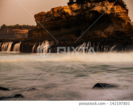 Long exposure view of silky sea water moving over dark rock terraces below rocky cliffs at Klayar Beach (Pantai Klayar), Pacitan, East Java, Indonesia. Long exposure view of silky sea water moving over dark rock terraces below rocky cliffs at Klayar Beach (Pantai Klayar), Pacitan, East Java, Indonesia. 142197801