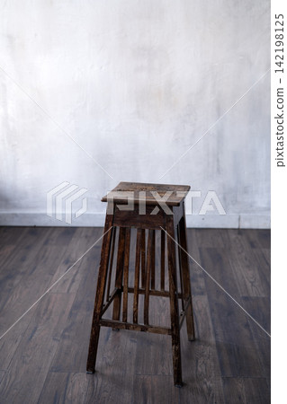 Vintage wooden stool standing on dark hardwood floor against rustic white concrete wall in minimal interior space Vintage wooden stool standing on dark hardwood floor against rustic white concrete wall in minimal interior space 142198125
