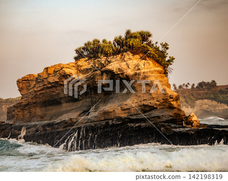 Distinct rocky hill formation with lush green vegetation rising from white ocean waves at Klayar Beach (Pantai Klayar), Pacitan, East Java, Indonesia. 142198319