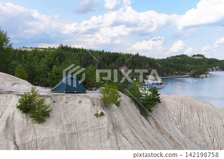 Camping tent on a rocky cliff overlooking a peaceful lake surrounded by forest under a blue sky with clouds, representing outdoor adventure, nature travel, recreation and wilderness. Camping tent on a rocky cliff overlooking a peaceful lake surrounded by forest under a blue sky with clouds, representing outdoor adventure, nature travel, recreation and wilderness. 142198758