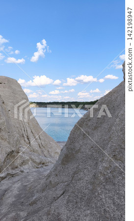 A turquoise lake with white rocky cliffs under a bright summer sky, a vertical picturesque natural backdrop, an idea for travel, relaxation and outdoor adventure. A turquoise lake with white rocky cliffs under a bright summer sky, a vertical picturesque natural backdrop, an idea for travel, relaxation and outdoor adventure. 142198947