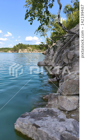 Turquoise quarry lake bordered by rugged rocky cliffs and green forest under a bright summer sky, with clear rippling water creating a scenic natural landscape for travel and outdoor concepts. Turquoise quarry lake bordered by rugged rocky cliffs and green forest under a bright summer sky, with clear rippling water creating a scenic natural landscape for travel and outdoor concepts. 142199487