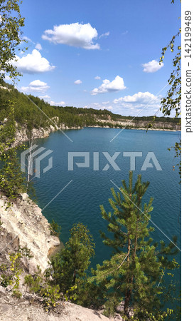 A picturesque blue quarry lake surrounded by trees, rocky cliffs, and lush forest under a summer blue sky. A vertical backdrop of natural landscapes, perfect for travel and outdoor recreation. A picturesque blue quarry lake surrounded by trees, rocky cliffs, and lush forest under a summer blue sky. A vertical backdrop of natural landscapes, perfect for travel and outdoor recreation. 142199489