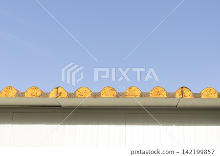 Corrugated roof edge with yellow tiles and white siding against blue sky Corrugated roof edge with yellow tiles and white siding against blue sky 142199857