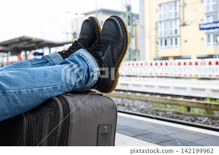 A person wearing jeans and black leather boots is resting with legs stretched over a suitcase while waiting for a train at a railway station platform. A person wearing jeans and black leather boots is resting with legs stretched over a suitcase while waiting for a train at a railway station platform. 142199962