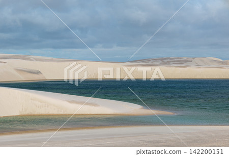 Beneath the blue-gray cloudy sky lies a deep blue lagoon and white sand dunes: Lençóis Maranhenses 142200151