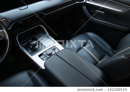 A wide armrest between the front seats trimmed with perforated leather inside a spacious car interior. Vehicle interior details. Armrest and leather seats in car A wide armrest between the front seats trimmed with perforated leather inside a spacious car interior. Vehicle interior details. Armrest and leather seats in car 142200554