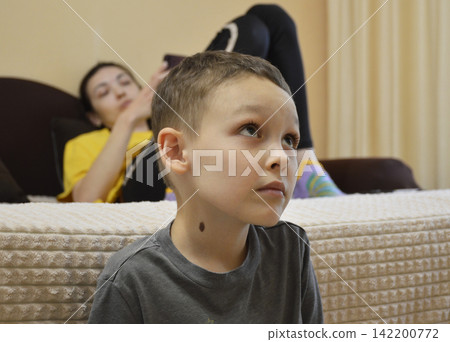 A young boy in a gray T-shirt is sitting on the edge of a bed or mattress. A young boy in a gray T-shirt is sitting on the edge of a bed or mattress. 142200772