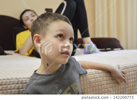 A young boy in a gray T-shirt is sitting on the edge of a bed or mattress. A young boy in a gray T-shirt is sitting on the edge of a bed or mattress. 142200773