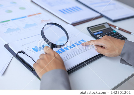 Financial expert closely inspects detailed corporate documents with magnifying glass at office desk. 142201201