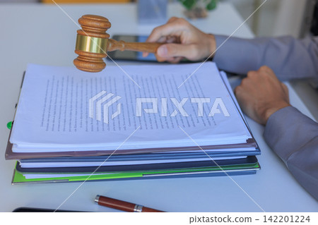 Legal professional hand holding a judge hammer over a stack of contracts on a desk. 142201224