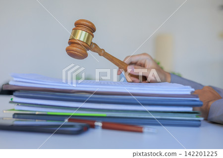 Legal professional hand holding a judge hammer over a stack of contracts on a desk. 142201225