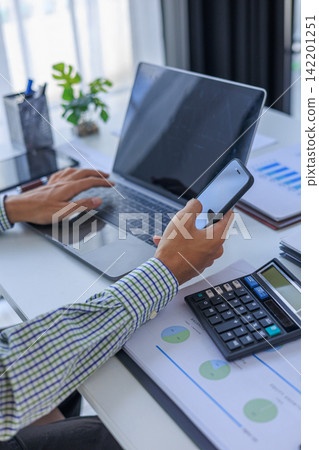 Businessman working in office using smartphone and typing on laptop with graph data and calculator at desk. Businessman working in office using smartphone and typing on laptop with graph data and calculator at desk. 142201251