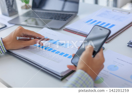 Professional financial analyst working on analyzing charts and documents using smartphone in modern office. Professional financial analyst working on analyzing charts and documents using smartphone in modern office. 142201259