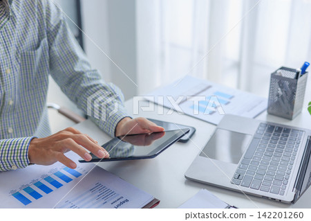 Professional financial analyst using tablet while working on analyzing charts and documents in modern office. Professional financial analyst using tablet while working on analyzing charts and documents in modern office. 142201260