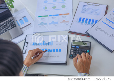 Close-up corporate accountant hand holding pen reviewing revenue growth charts and budget document in bright office. 142201269
