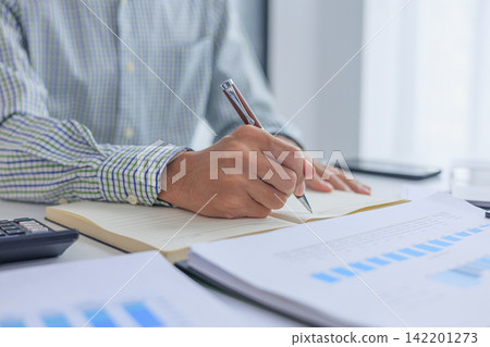 Businessman writing in notebook while working on analyzing financial report data graph chart at desk in office. Businessman writing in notebook while working on analyzing financial report data graph chart at desk in office. 142201273