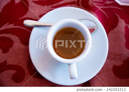 espresso cup resting on saucer atop red patterned cloth in relaxed coffee culture setting top-down view with ceramic and metallic materials white and brown tones. espresso cup resting on saucer atop red patterned cloth in relaxed coffee culture setting top-down view with ceramic and metallic materials white and brown tones. 142201511