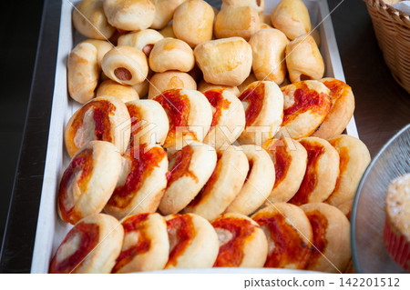 Tray of assorted baked bread rolls with golden crust and red sauce topping, arranged in close packed rows for bakery display, buffet service, snack table, casual catering, and fresh food presentation Tray of assorted baked bread rolls with golden crust and red sauce topping, arranged in close packed rows for bakery display, buffet service, snack table, casual catering, and fresh food presentation 142201512