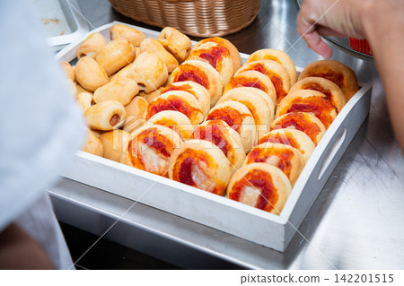 Freshly baked stuffed bread rolls with tomato filling on stainless steel counter beside reaching hand choosing snacks for buffet service in busy commercial kitchen 142201515