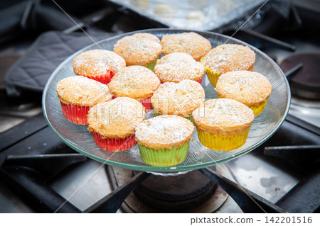 Powdered sugar cupcakes in colorful paper liners arranged inside clear glass bowl above stovetop burners, showing homemade dessert presentation with soft golden tops and cozy kitchen warmth 142201516