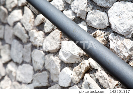 Black cable running diagonally across rough white stone wall with shallow depth of field and bright daylight creating strong contrast between smooth industrial surface and textured masonry Black cable running diagonally across rough white stone wall with shallow depth of field and bright daylight creating strong contrast between smooth industrial surface and textured masonry 142201517