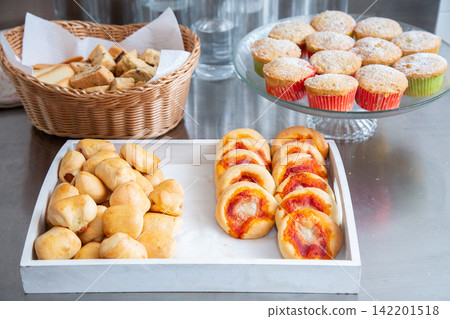 Assorted freshly baked pastries, muffins, rolls, cookies and mini pizzas arranged on serving trays and wicker basket at buffet table with warm golden tones and casual catering presentation 142201518