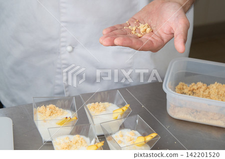 Chef hand sprinkling crumb topping over individual dessert cups with creamy layers and fruit garnish on kitchen counter during pastry preparation inside bright modern kitchen Chef hand sprinkling crumb topping over individual dessert cups with creamy layers and fruit garnish on kitchen counter during pastry preparation inside bright modern kitchen 142201520