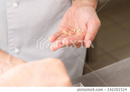 Close-up of hands holding small crumbs over kitchen counter beside white chef coat, showing food preparation detail, careful handling, and soft indoor light with shallow focus Close-up of hands holding small crumbs over kitchen counter beside white chef coat, showing food preparation detail, careful handling, and soft indoor light with shallow focus 142201524