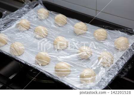 Fresh dough balls resting on lined baking tray under clear plastic wrap, arranged for proofing in home kitchen with soft light, clean surfaces, and preparation for baking Fresh dough balls resting on lined baking tray under clear plastic wrap, arranged for proofing in home kitchen with soft light, clean surfaces, and preparation for baking 142201528