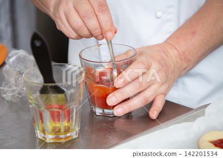 Hands stirring red sauce in glass with spoon beside oil glass on stainless kitchen counter, close view of culinary preparation by person wearing white chef coat in bright restaurant workspace Hands stirring red sauce in glass with spoon beside oil glass on stainless kitchen counter, close view of culinary preparation by person wearing white chef coat in bright restaurant workspace 142201534