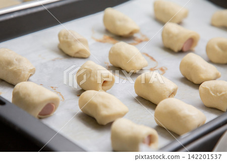 Raw dough wrapped around small sausage pieces on parchment lined baking tray, arranged for baking with soft kitchen light, close up food preparation scene with simple homemade snack making Raw dough wrapped around small sausage pieces on parchment lined baking tray, arranged for baking with soft kitchen light, close up food preparation scene with simple homemade snack making 142201537