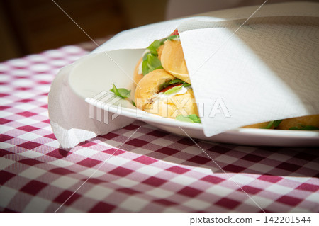 Fresh bread rolls on white plate beneath napkin, warm sunlight over red and white checkered tablecloth creating rustic breakfast dining mood in cozy indoor setting Fresh bread rolls on white plate beneath napkin, warm sunlight over red and white checkered tablecloth creating rustic breakfast dining mood in cozy indoor setting 142201544