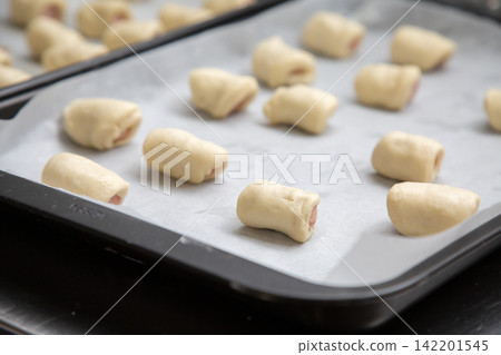 Unbaked pastry rolls arranged on parchment lined baking tray, showing soft dough pieces with filling ends, ready for oven in close up kitchen preparation still life 142201545