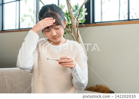 A primary school or junior high school girl sitting on a sofa at home feeling unwell and taking her temperature with a thermometer 142202010