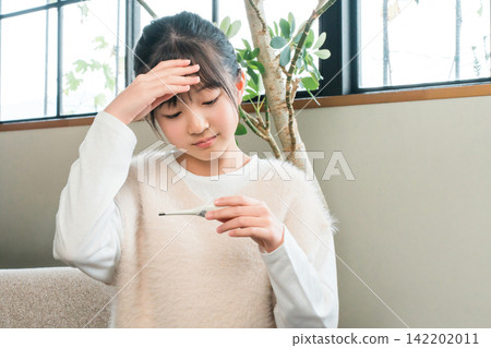 A primary school or junior high school girl sitting on a sofa at home feeling unwell and taking her temperature with a thermometer A primary school or junior high school girl sitting on a sofa at home feeling unwell and taking her temperature with a thermometer 142202011