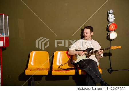 Friendly musician calmly strumming guitar on train station seats Friendly musician calmly strumming guitar on train station seats 142202611
