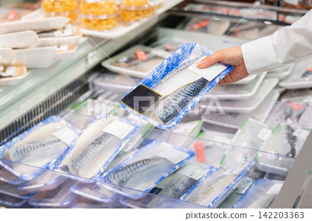 A woman picks up a pack of mackerel fillets and chooses one at the fresh fish counter of a supermarket. 142203363