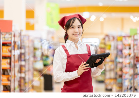 A female store clerk at the supermarket is working with a smile on her face while holding a tablet. A female store clerk at the supermarket is working with a smile on her face while holding a tablet. 142203379