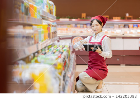 A female employee in the beverage section of a supermarket is holding a tablet to check products. A female employee in the beverage section of a supermarket is holding a tablet to check products. 142203388