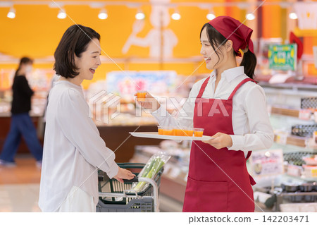 A smiling female staff member offers a free sample drink to a female customer at a supermarket. A smiling female staff member offers a free sample drink to a female customer at a supermarket. 142203471