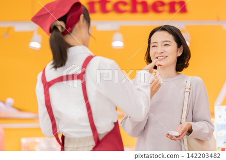 一位微笑的女顧客正在超市品嚐樣品,一位女店員正在協助她。 一位微笑的女顧客正在超市品嚐樣品,一位女店員正在協助她。 142203482