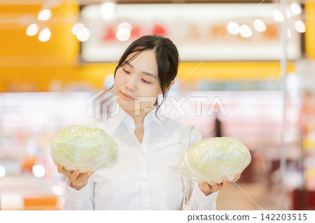 A young woman compares and chooses between two cabbages in the vegetable section of a supermarket. A young woman compares and chooses between two cabbages in the vegetable section of a supermarket. 142203515